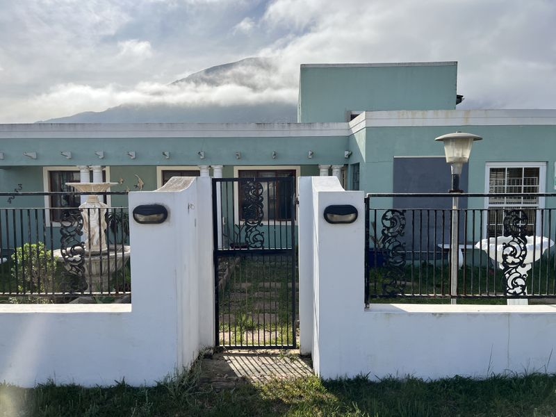 Front elevation of a house featuring a gated entrance, masonry boundary wall, and decorative fountain with mountain views in the background.