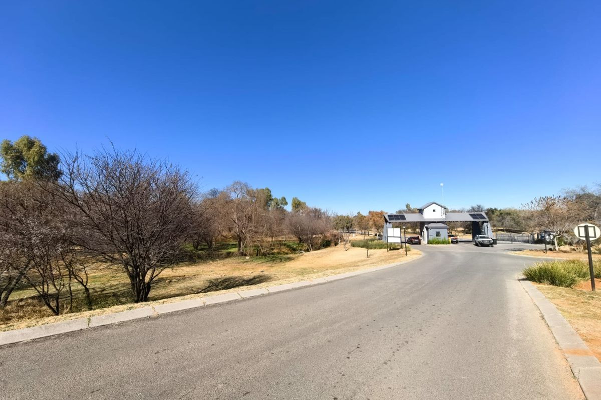 A view of the residential estate entrance featuring an asphalt road leading to a security gatehouse equipped with solar panels.
