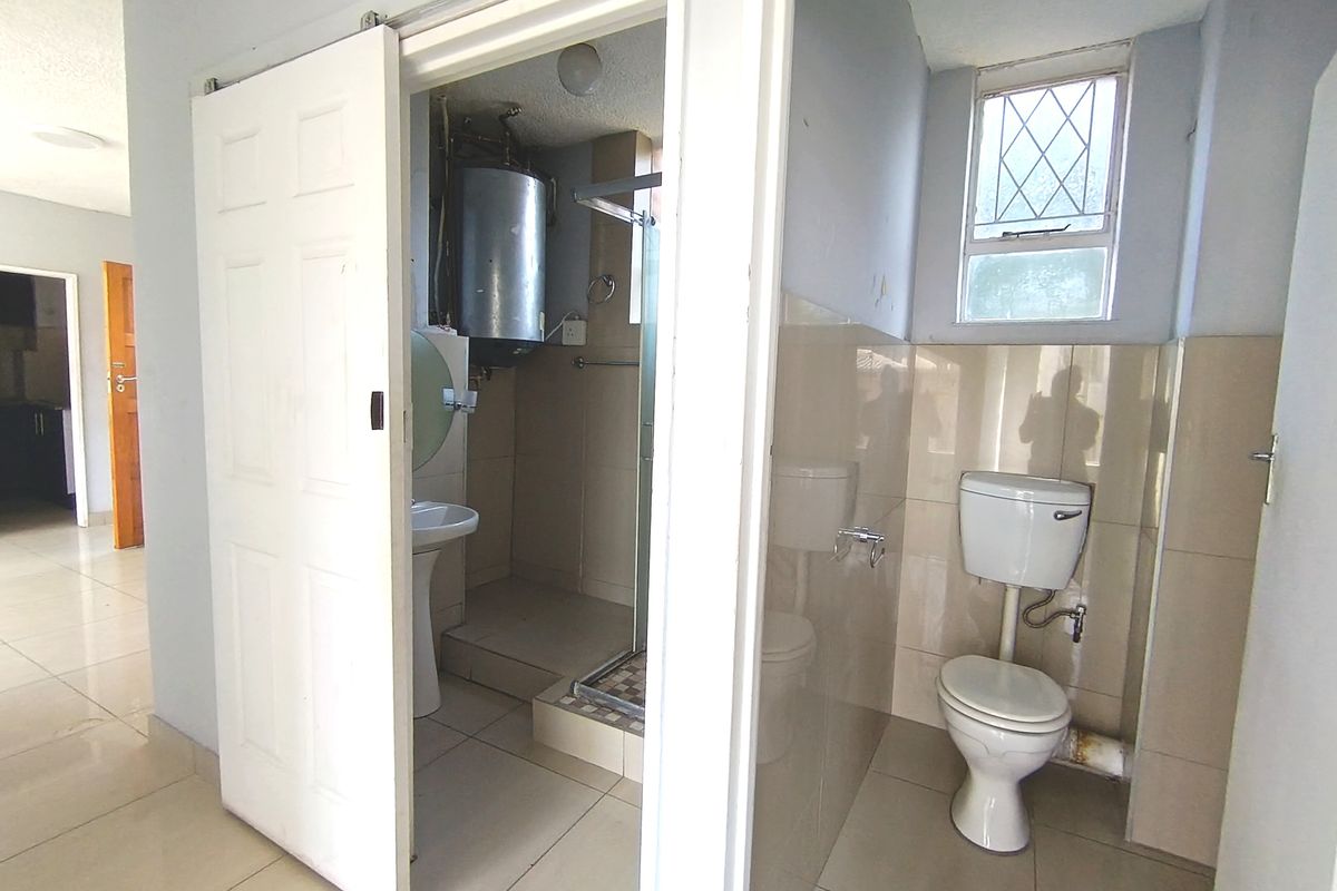A functional bathroom featuring a pedestal basin, shower enclosure, and toilet, with tiled finishes and an exposed electric geyser.
