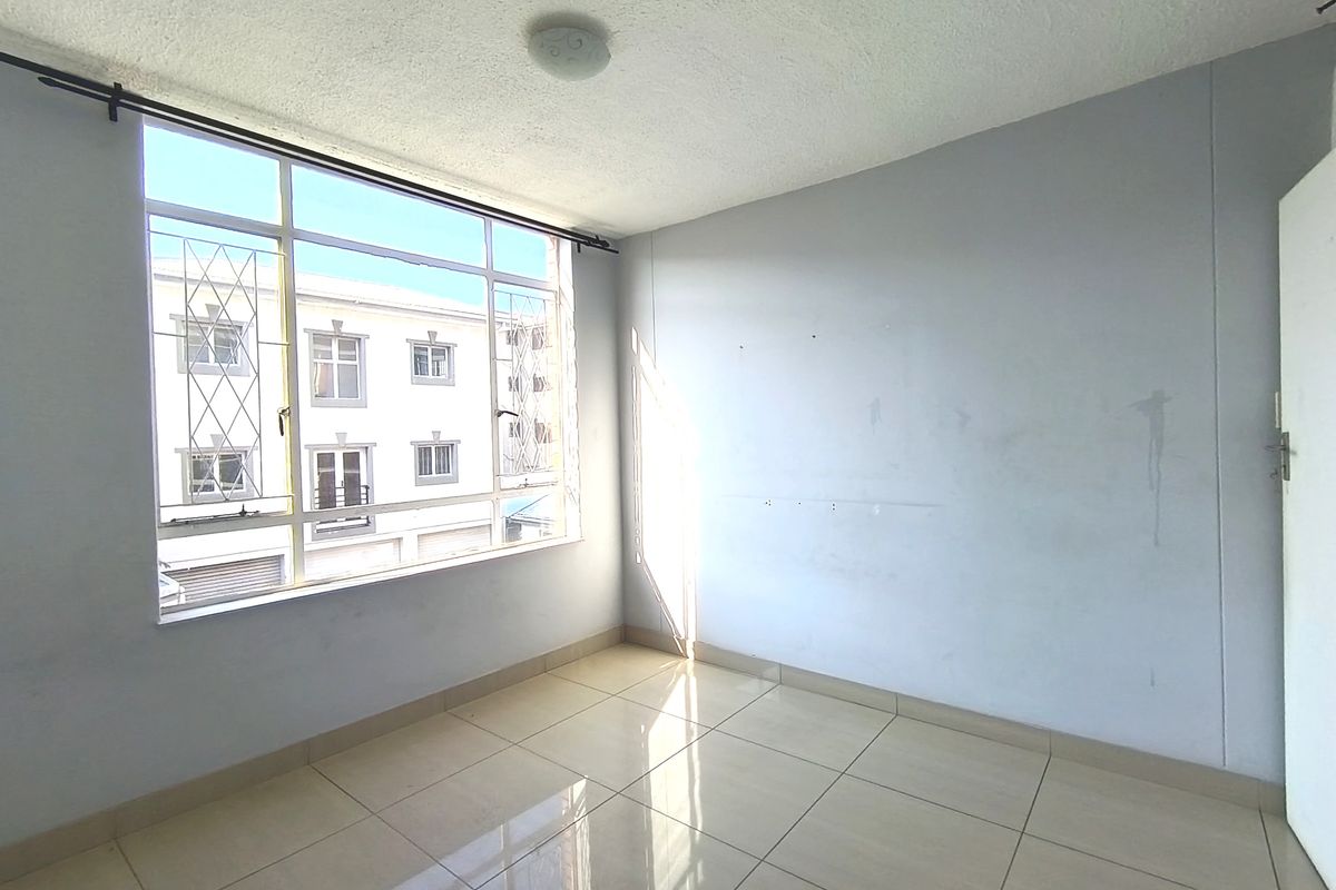 An empty bedroom featuring tiled flooring, a large window with security bars, and plain walls in need of cosmetic attention.