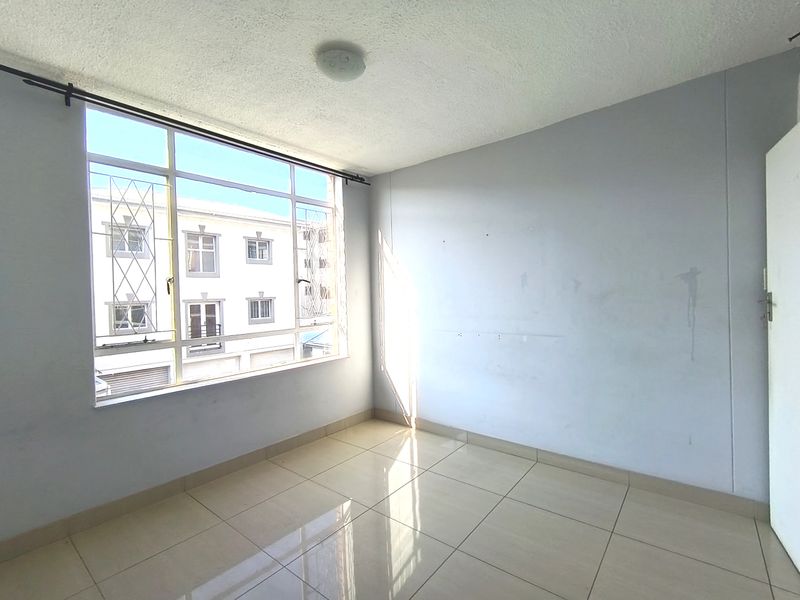 An empty bedroom featuring tiled flooring, a large window with security bars, and plain walls in need of cosmetic attention.