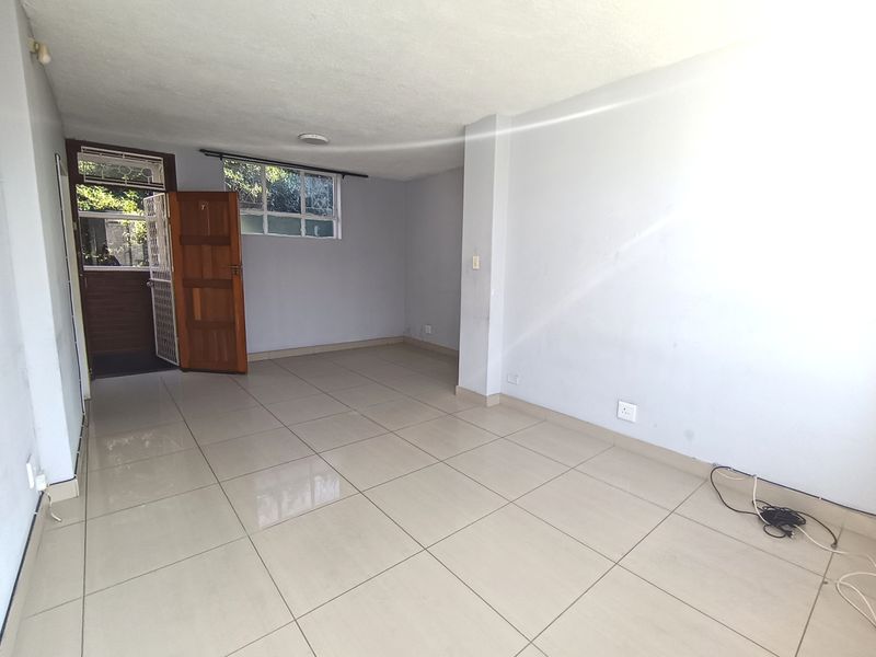 An empty, open-plan living area featuring neutral tiled flooring, a timber entrance door with a security gate, and windows with security bars.