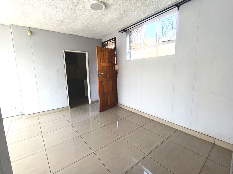 An empty living room featuring neutral tiled flooring, a timber entrance door with security gate, and a window with integrated burglar bars.