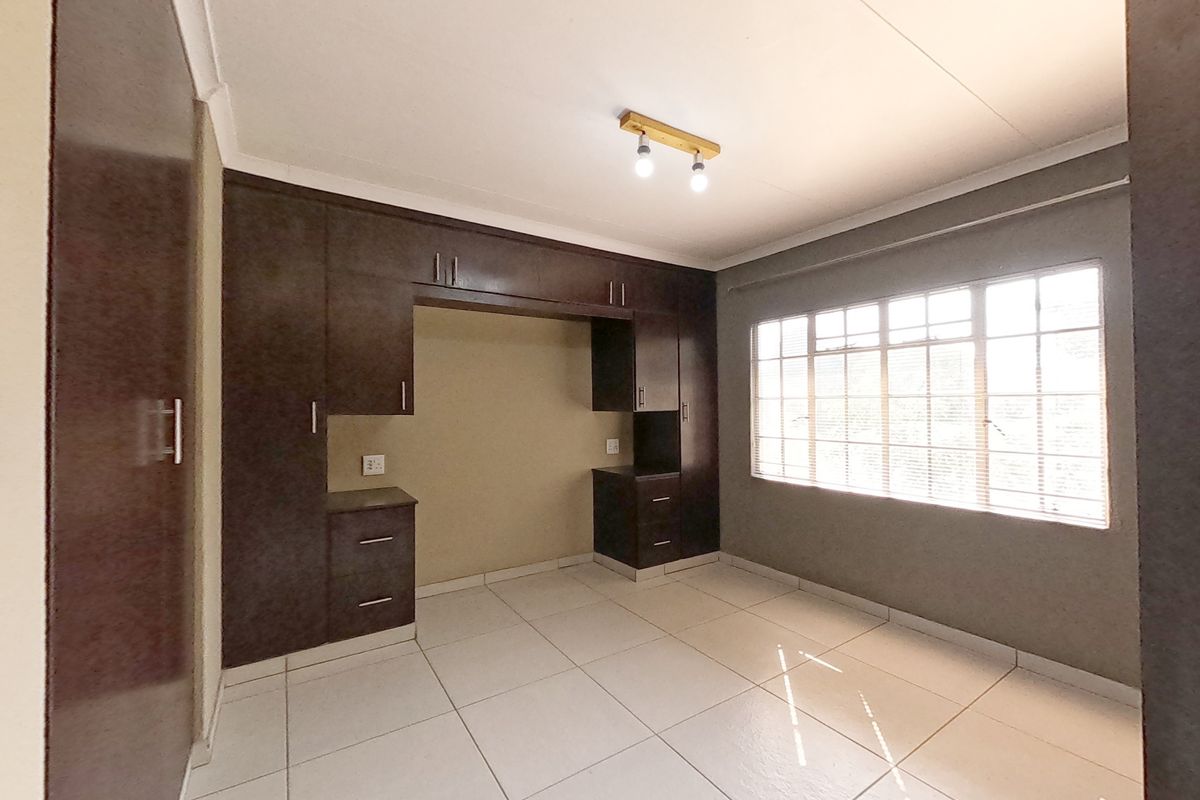 A bright, empty bedroom featuring neutral tiled flooring, built-in dark wood cabinetry, and a large window with security bars.
