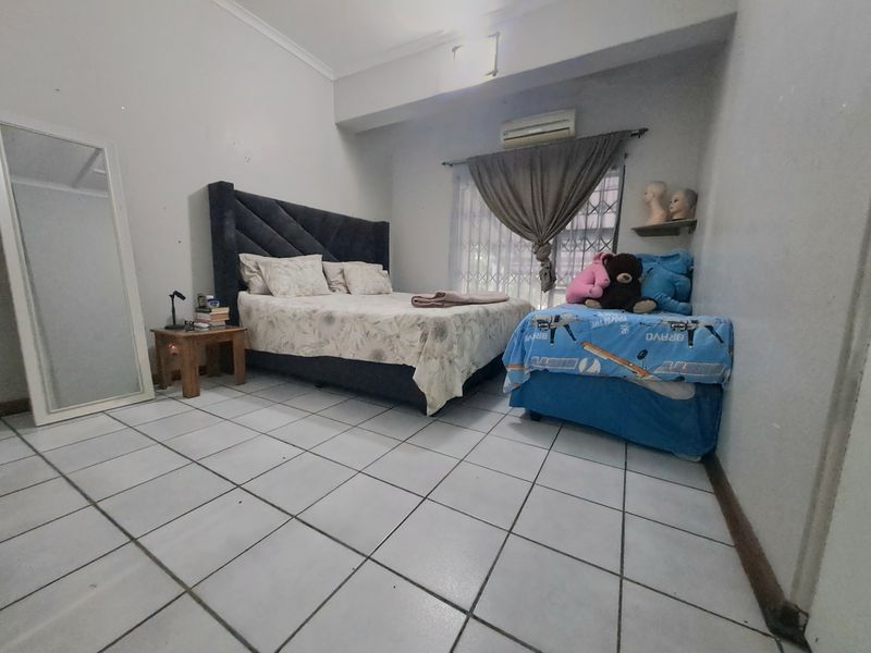 A functional bedroom featuring tiled flooring, a wall-mounted air conditioning unit, and security gate access to the window.