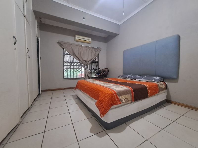 A bedroom featuring tiled flooring, built-in cupboards, and an air conditioning unit, with security bars installed on the window.