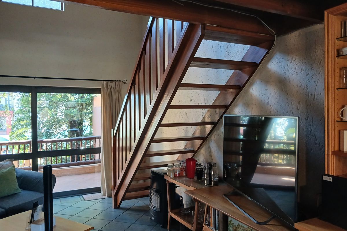 A living room featuring a prominent wooden staircase, exposed timber ceiling beams, tiled flooring, and a sliding glass door leading to an outdoor balcony.