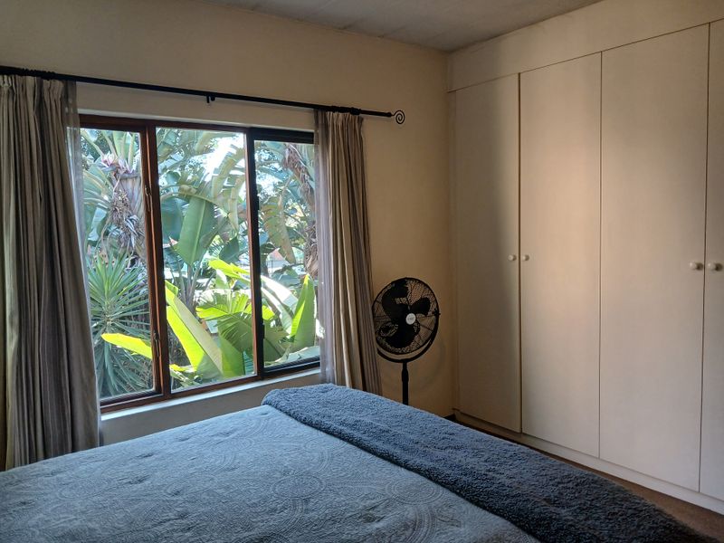 A bedroom featuring built-in cupboards and a large window overlooking lush garden greenery.