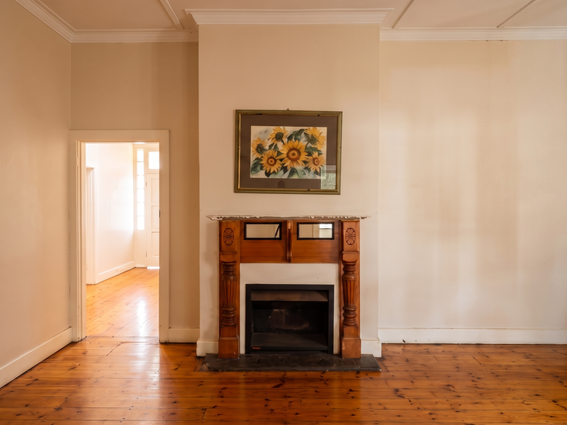 A spacious living room featuring original timber flooring, a classic wooden fireplace mantel, and crown molding, offering a bright and neutral canvas.
