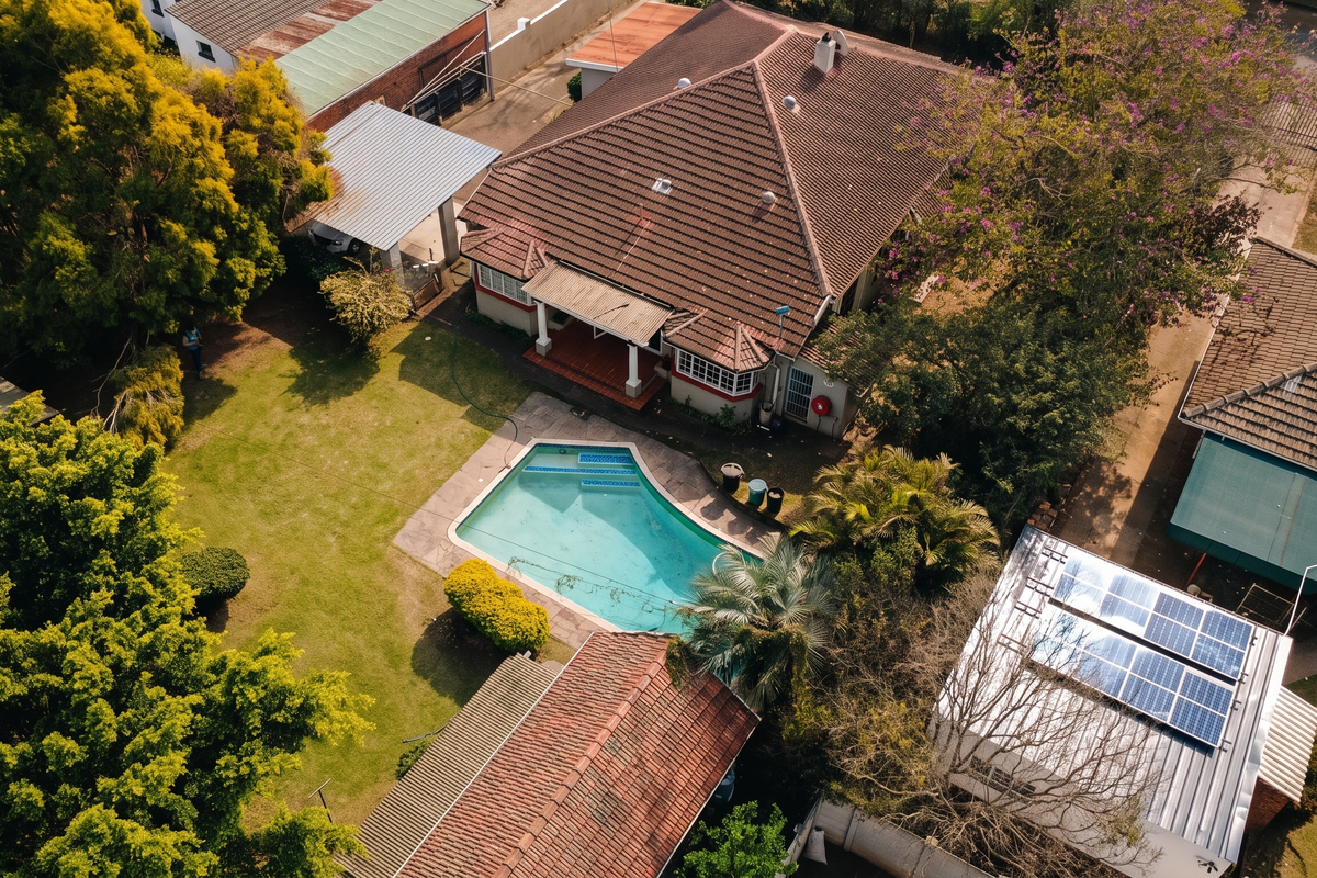 Aerial view of a residential property featuring a swimming pool, mature garden, tiled roof, and solar panels installed on an outbuilding.