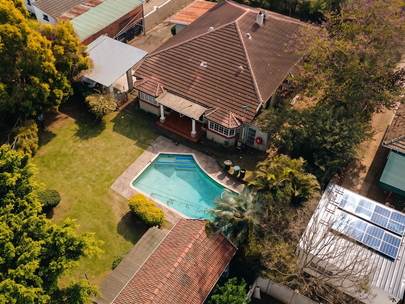 Aerial view of a residential property featuring a swimming pool, mature garden, tiled roof, and solar panels installed on an outbuilding.