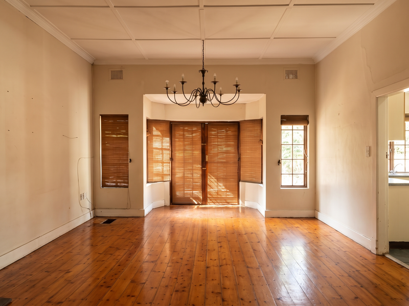 Spacious living room featuring original hardwood flooring, a classic fireplace mantel, and a bay window with natural light, offering a blank canvas for renovation.