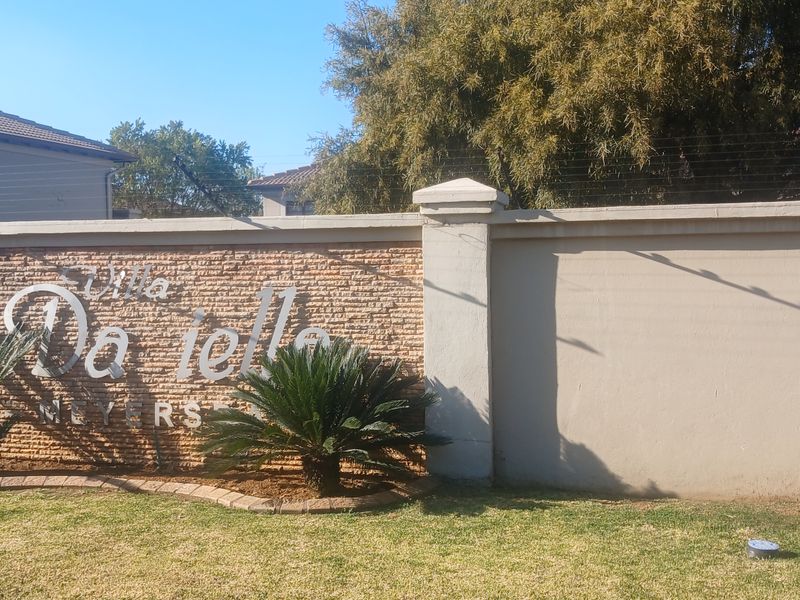 The front entrance area of the Villa Da Ielle complex, featuring a stone-clad signage wall, perimeter security fencing, and manicured landscaping.
