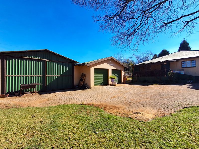 Rear exterior view featuring a paved driveway, a double garage, and a large corrugated metal storage shed set against a clear blue sky.