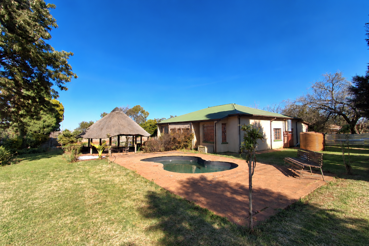 Rear garden view featuring a swimming pool, brick paved patio area, and a traditional thatched lapa structure set within a spacious lawn.