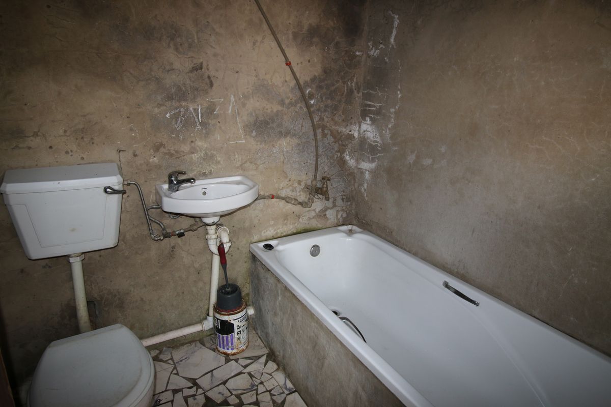 A dilapidated bathroom featuring a basic bathtub, toilet, and wall-mounted basin, requiring significant renovation and surface repairs to restore functionality.