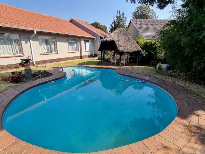 A well-maintained backyard featuring a curved swimming pool with brick paving and a traditional thatched lapa for outdoor entertainment.