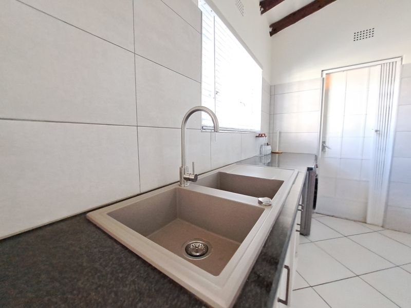 A clean, modern kitchen area featuring a double basin sink, granite countertops, and neutral ceramic tiling with exposed timber rafters.