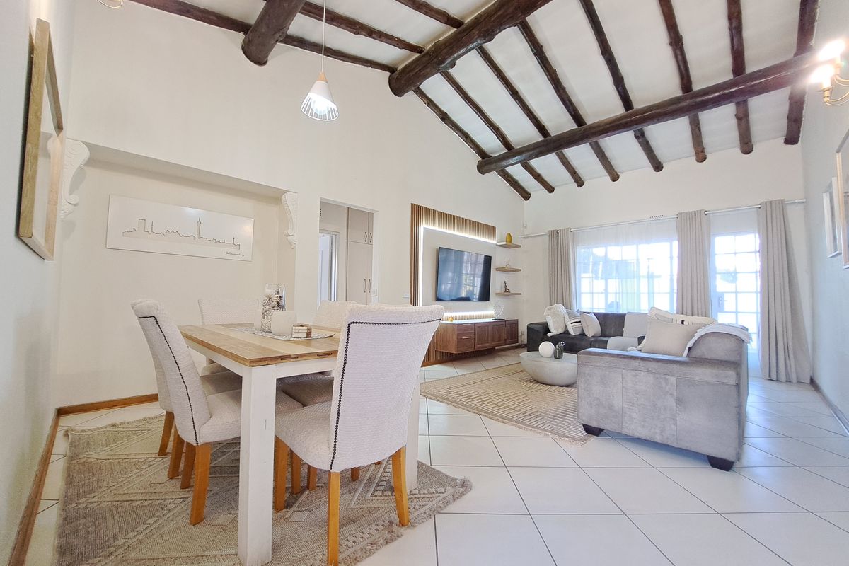 Spacious open-plan living and dining area featuring high vaulted ceilings with exposed timber beams, tiled flooring, and a modern built-in media wall.