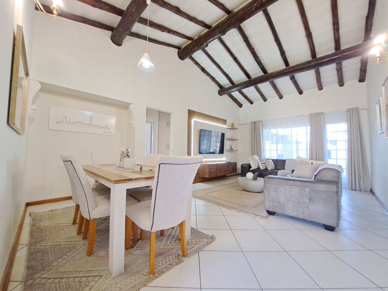 Spacious open-plan living and dining area featuring high vaulted ceilings with exposed timber beams, tiled flooring, and a modern built-in media wall.
