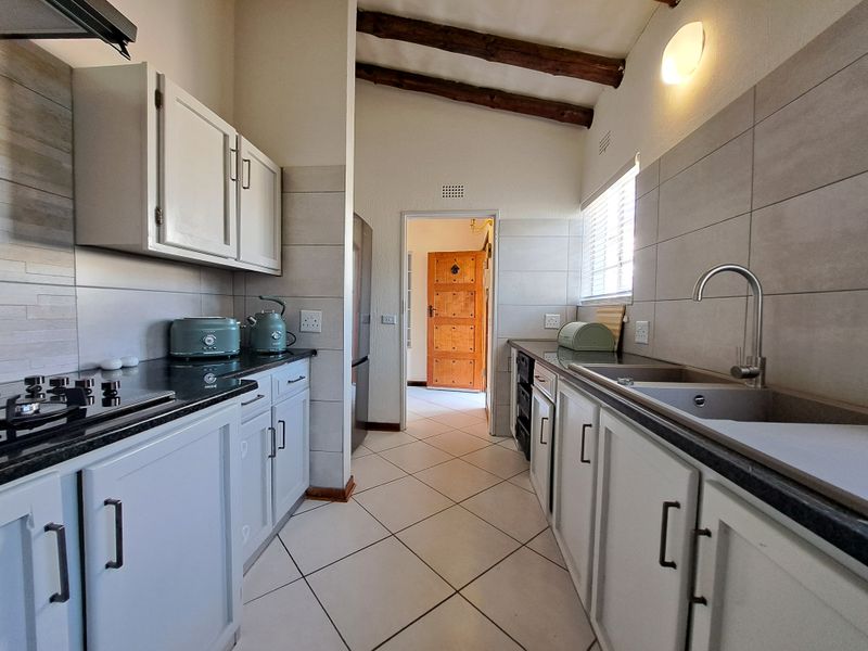 A functional galley-style kitchen featuring granite countertops, built-in cabinetry, a gas hob, and exposed timber ceiling beams, leading to an adjacent room.