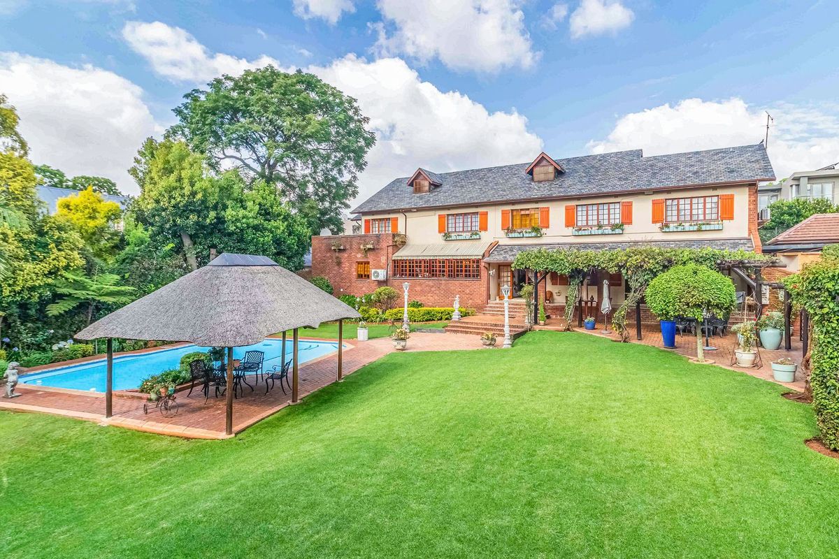 Spacious rear garden featuring a swimming pool, thatched gazebo, brick patio, and a well-maintained lawn with mature landscaping and a charming brick residence.