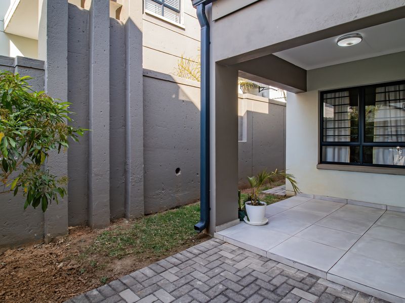 A covered patio area featuring tiled flooring, a downpipe, and security-barred windows, adjacent to a paved walkway and concrete boundary wall.