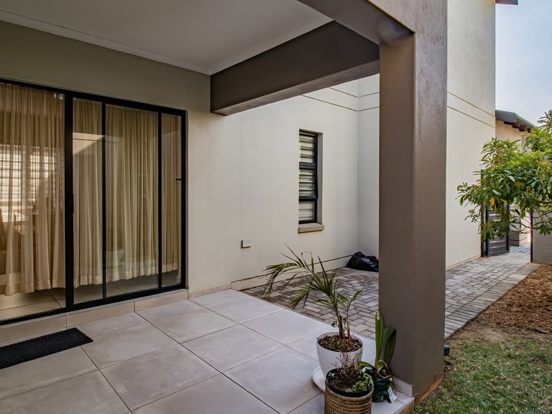 A covered patio area featuring tiled flooring, a sliding glass door, and a paved walkway leading into the garden space.