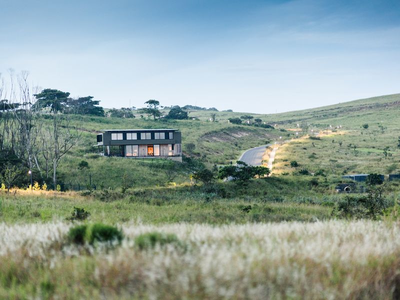 A scenic landscape view featuring a modern residential home nestled within a vast, grassy hillside setting with an adjacent paved road.