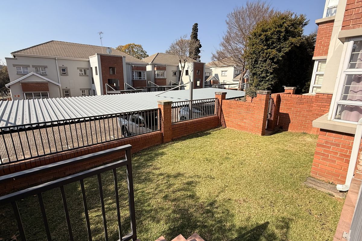 A private rear garden area featuring a well-maintained lawn, enclosed by brick boundary walls and metal security fencing, overlooking a communal carport area.