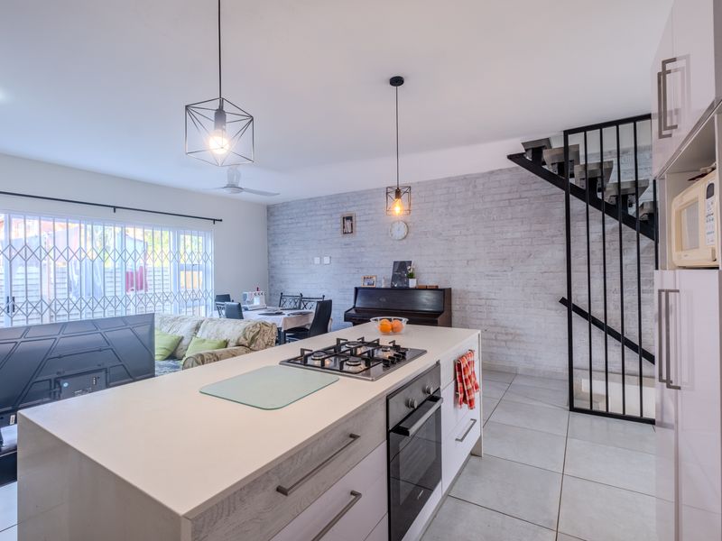 Modern open-plan living area featuring a kitchen island with gas hob, exposed brick wall, and a contemporary metal staircase.