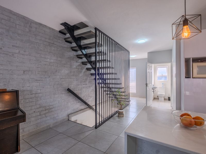 Modern open-plan living area featuring an industrial-style metal staircase, exposed brick wall, and sleek tiled flooring, leading to a guest bathroom.