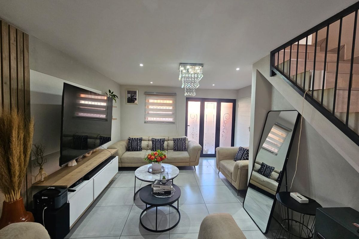 A well-maintained living room featuring large format floor tiles, modern recessed lighting, a decorative crystal chandelier, and an open staircase with a metal balustrade.