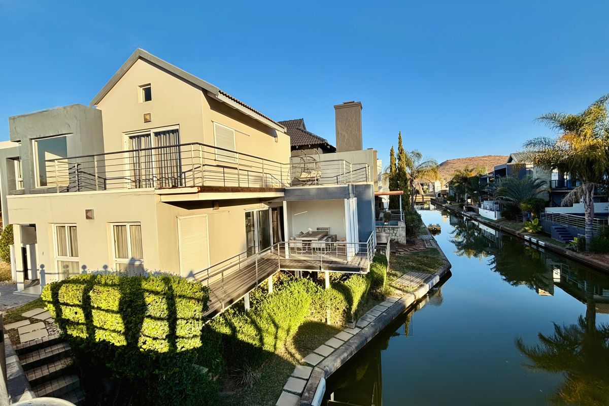 Rear view of a multi-level house featuring a balcony, covered patio, and direct access to a canal, set within a landscaped estate environment.