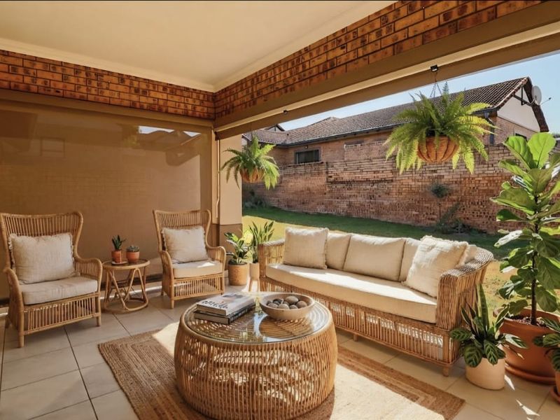 A covered outdoor patio featuring exposed brick walls, tiled flooring, and retractable blinds, overlooking a private garden area.