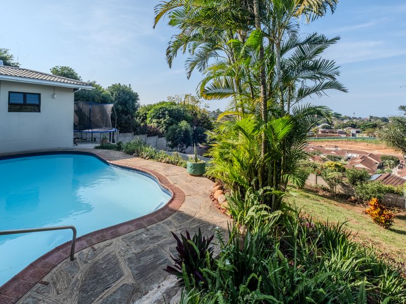 A well-maintained swimming pool area featuring a stone-paved deck, landscaped garden, and scenic views of the surrounding neighborhood.