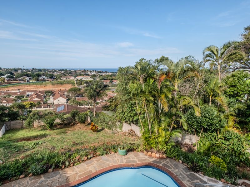 An elevated view of a swimming pool with stone paving, overlooking a lush garden and expansive suburban landscape with distant sea views.