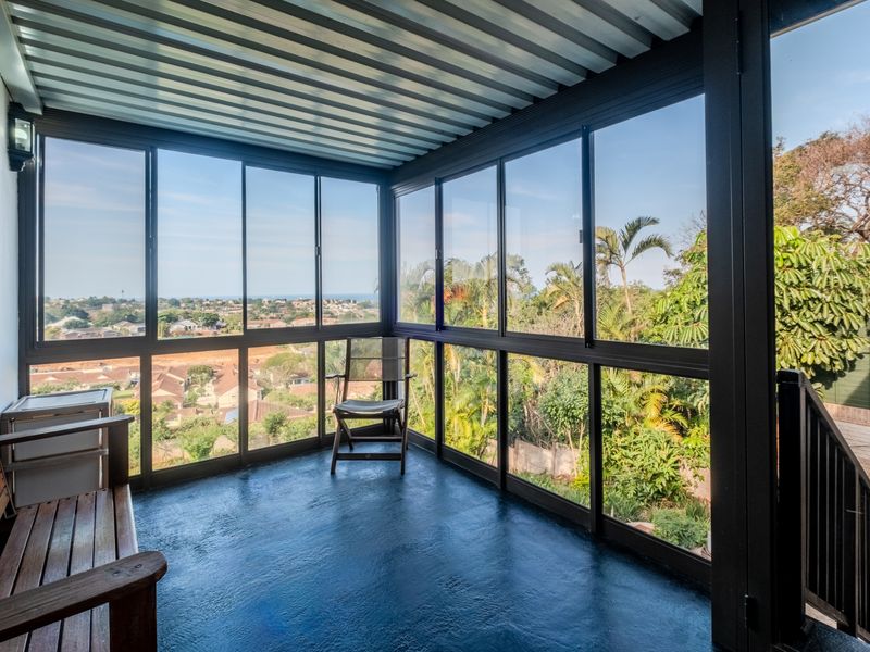 An enclosed balcony featuring large aluminum-framed windows, a corrugated metal ceiling, and a painted concrete floor, offering elevated views of the surrounding neighborhood.