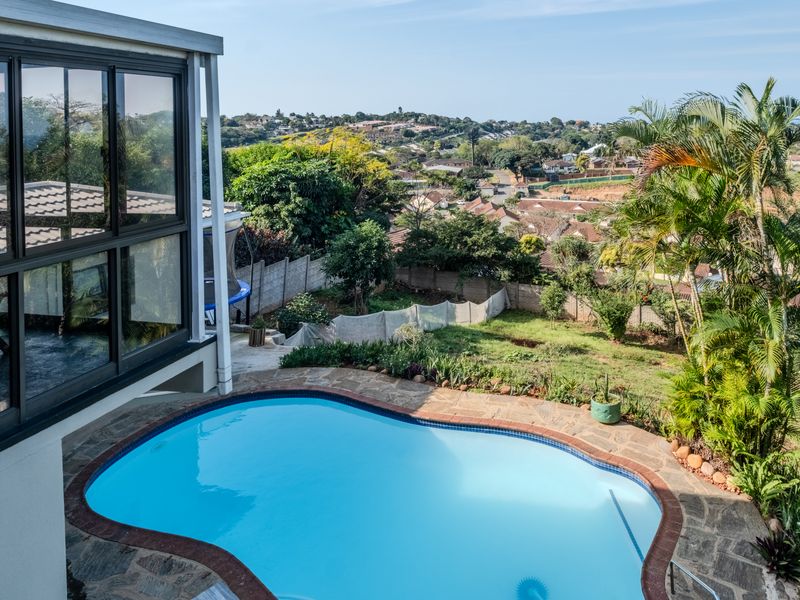 An elevated view of a swimming pool with stone paving, overlooking a residential garden and surrounding neighbourhood.