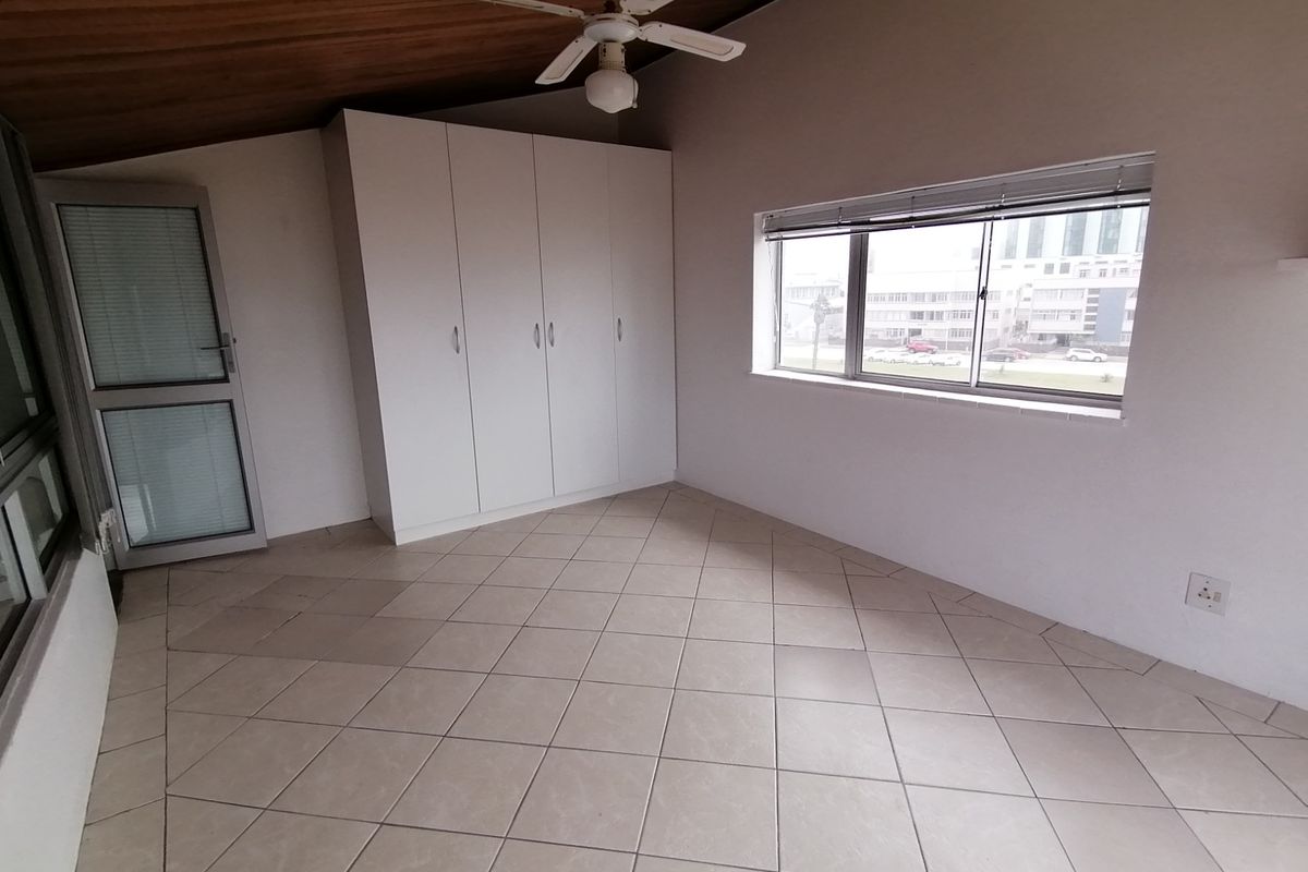 A bright, empty bedroom featuring tiled flooring, built-in cupboards, a ceiling fan, and a timber-clad ceiling with views of the surrounding area.