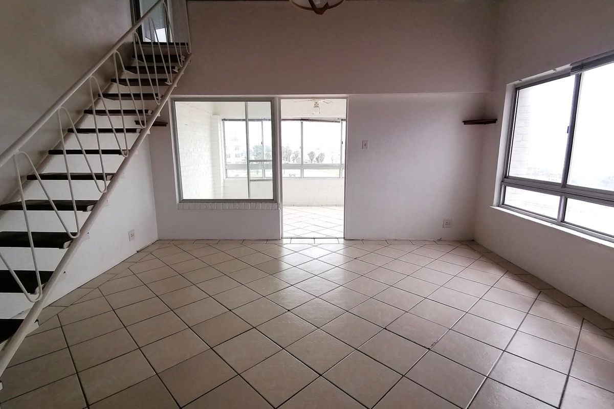 An empty, open-plan living area featuring tiled floors, a metal staircase leading to an upper level, and large windows providing natural light.
