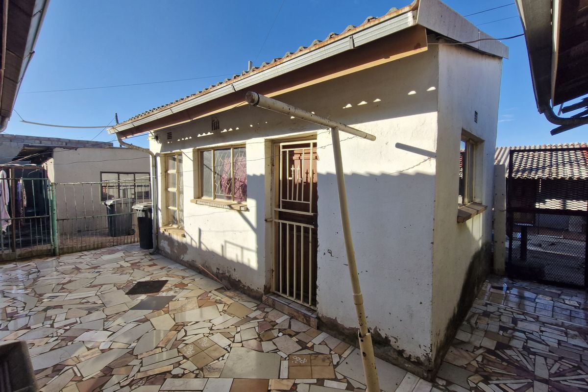 Rear exterior view of a small outbuilding featuring a tiled patio area, security gate, and metal window frames, showing signs of exterior wear.