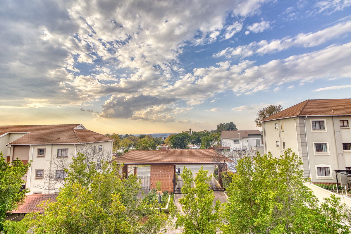 An elevated view of the apartment complex entrance, featuring a brick security gatehouse, paved driveway, and well-maintained residential buildings with tiled roofs.