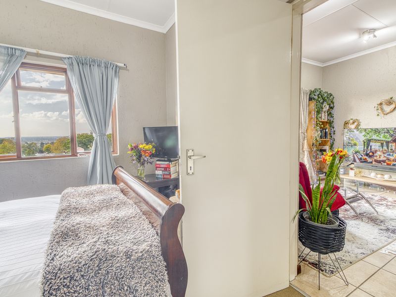 A bedroom featuring a window with exterior views, leading into an adjacent living area with tiled flooring and coved ceilings.
