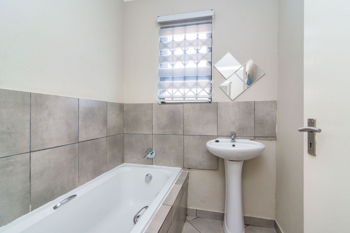 A functional bathroom featuring a pedestal basin, built-in bathtub, and neutral grey wall tiling with a roller blind for privacy.