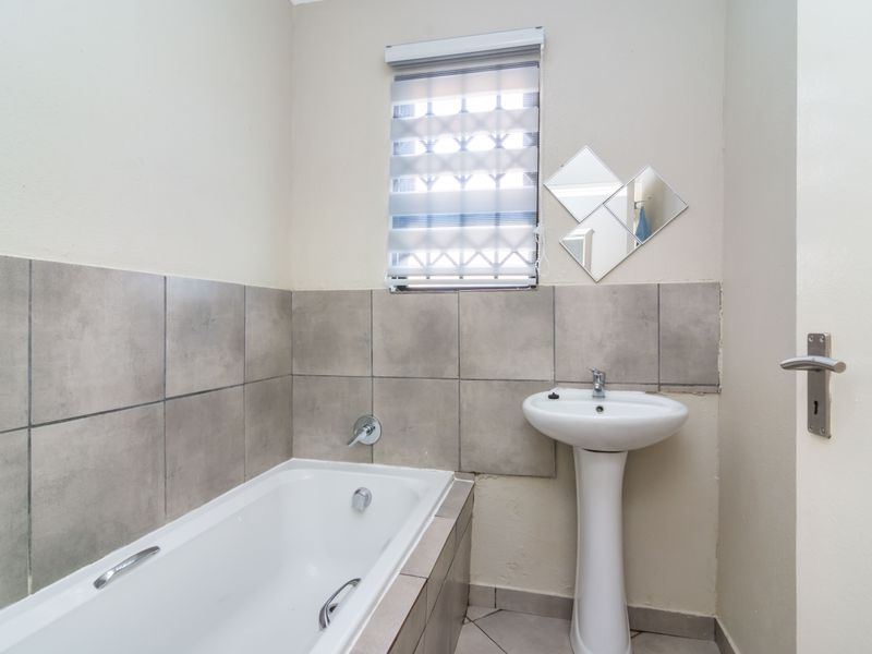 A functional bathroom featuring a pedestal basin, built-in bathtub, and neutral grey wall tiling with a roller blind for privacy.