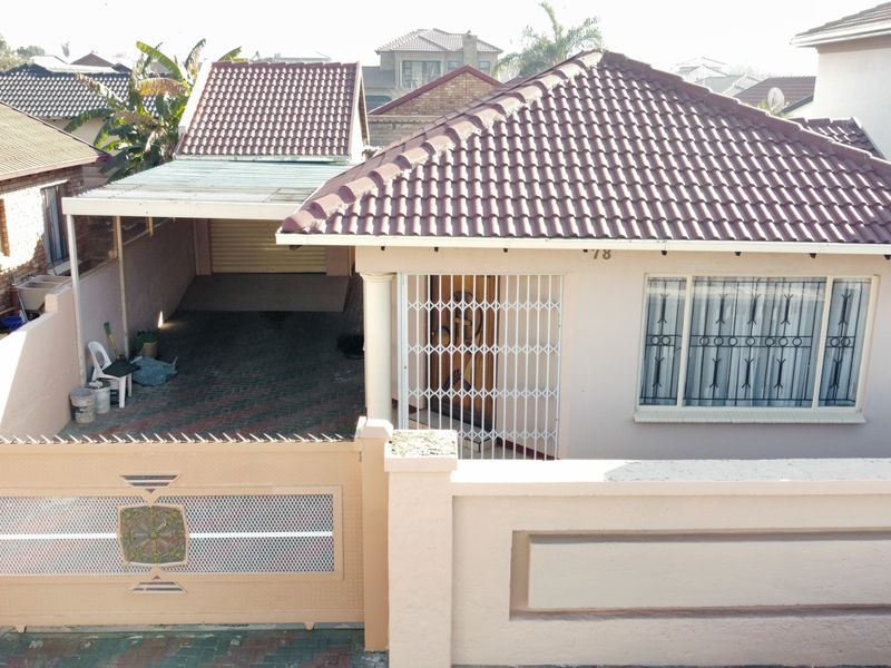 Front elevation of a residential house featuring a tiled roof, paved driveway, carport, and secure boundary wall with a metal gate.