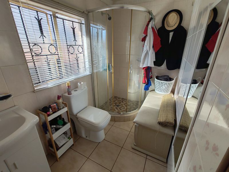 Compact bathroom featuring a corner shower enclosure, tiled walls and flooring, a standard toilet, and a vanity basin with security bars on the window.