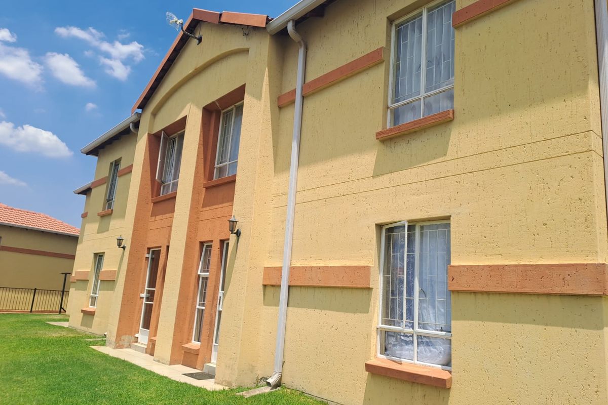 A side view of a two-story townhouse featuring textured yellow and orange facade, tiled roofing, and security-fitted windows overlooking a lawn.