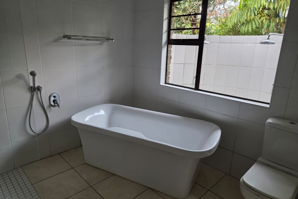 A clean, modern bathroom featuring a freestanding bathtub, a toilet, and a shower area with tiled finishes and a window overlooking the garden.